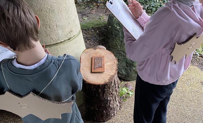 Boy and girl writing on clipboards as part of their outdoor activity. They are also wearing cardboard bats on their back.